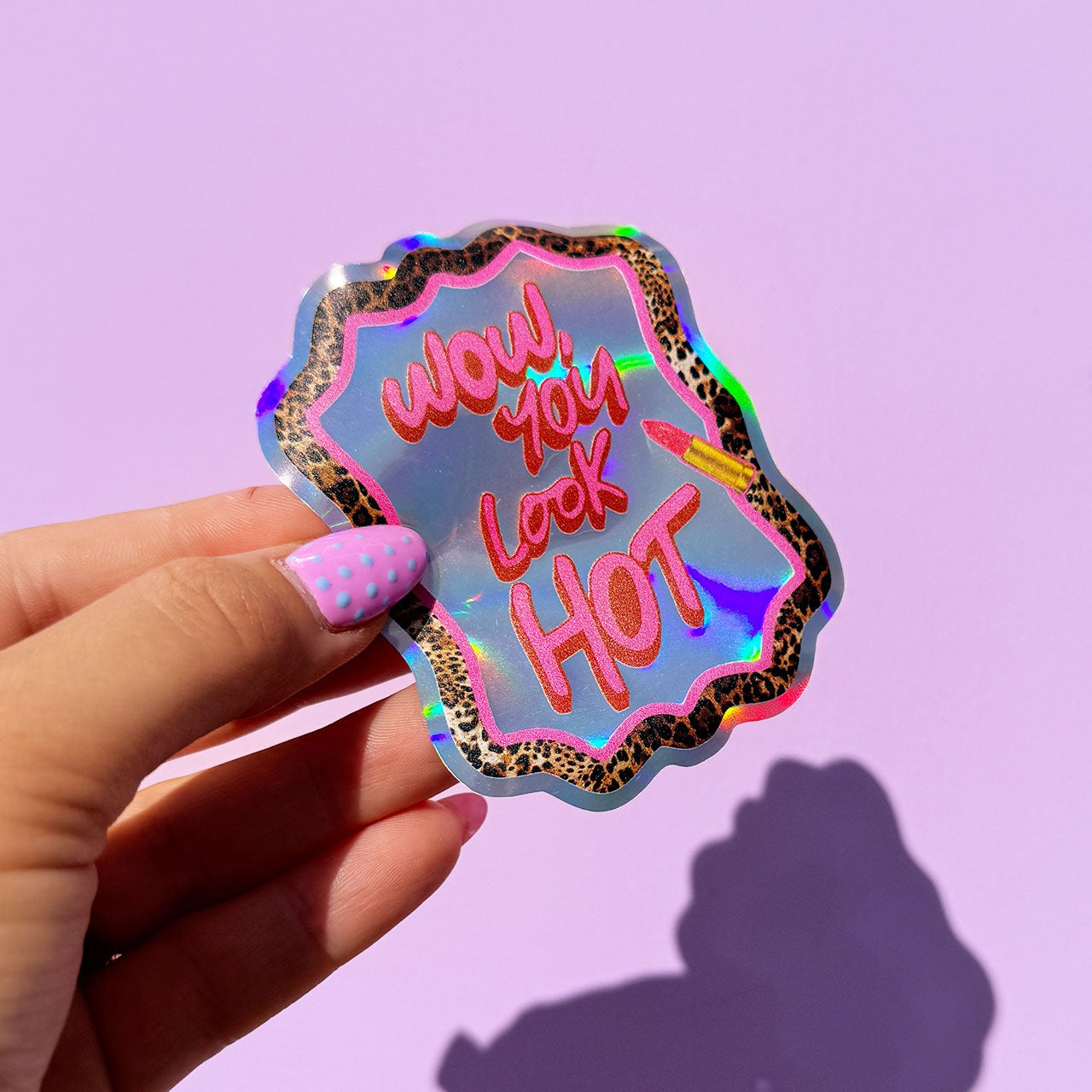 Holographic sticker with 'Wow you look hot' text held by a hand against a purple background