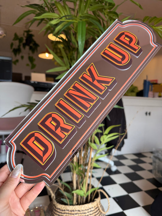 A person is holding a sign that says "DRINK UP" in a room with plants and a checkered floor.