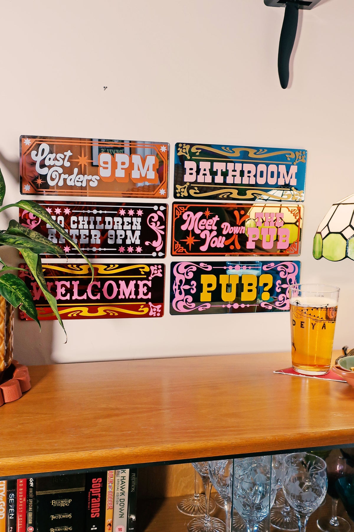 a wall with colorful, decorative signs advertising various establishments, including a pub, a children's play area, and a bathroom. There is also a glass of beer on a table in the foreground.