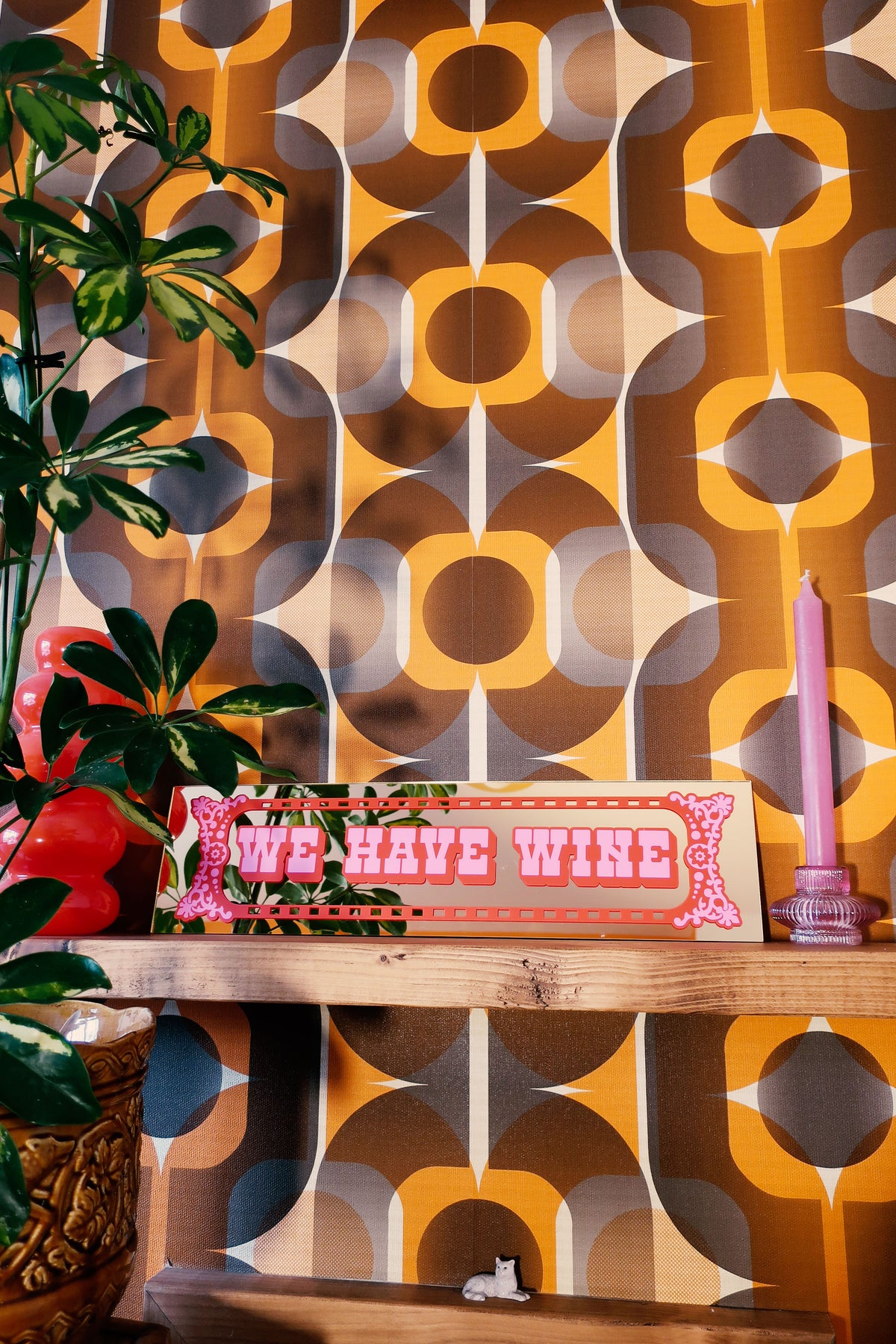 a shelf with a sign that reads "We have wine", a pink candle, and a plant. The background has a patterned wallpaper with a repeating design of circles and lines in shades of orange, brown, and gray.