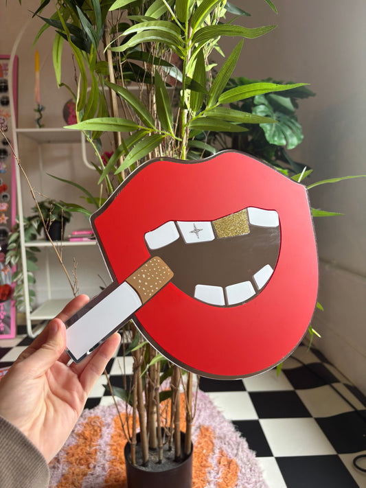 A hand holding a red and white sign with a graphic of a mouth and a cigarette, with a plant in the background.