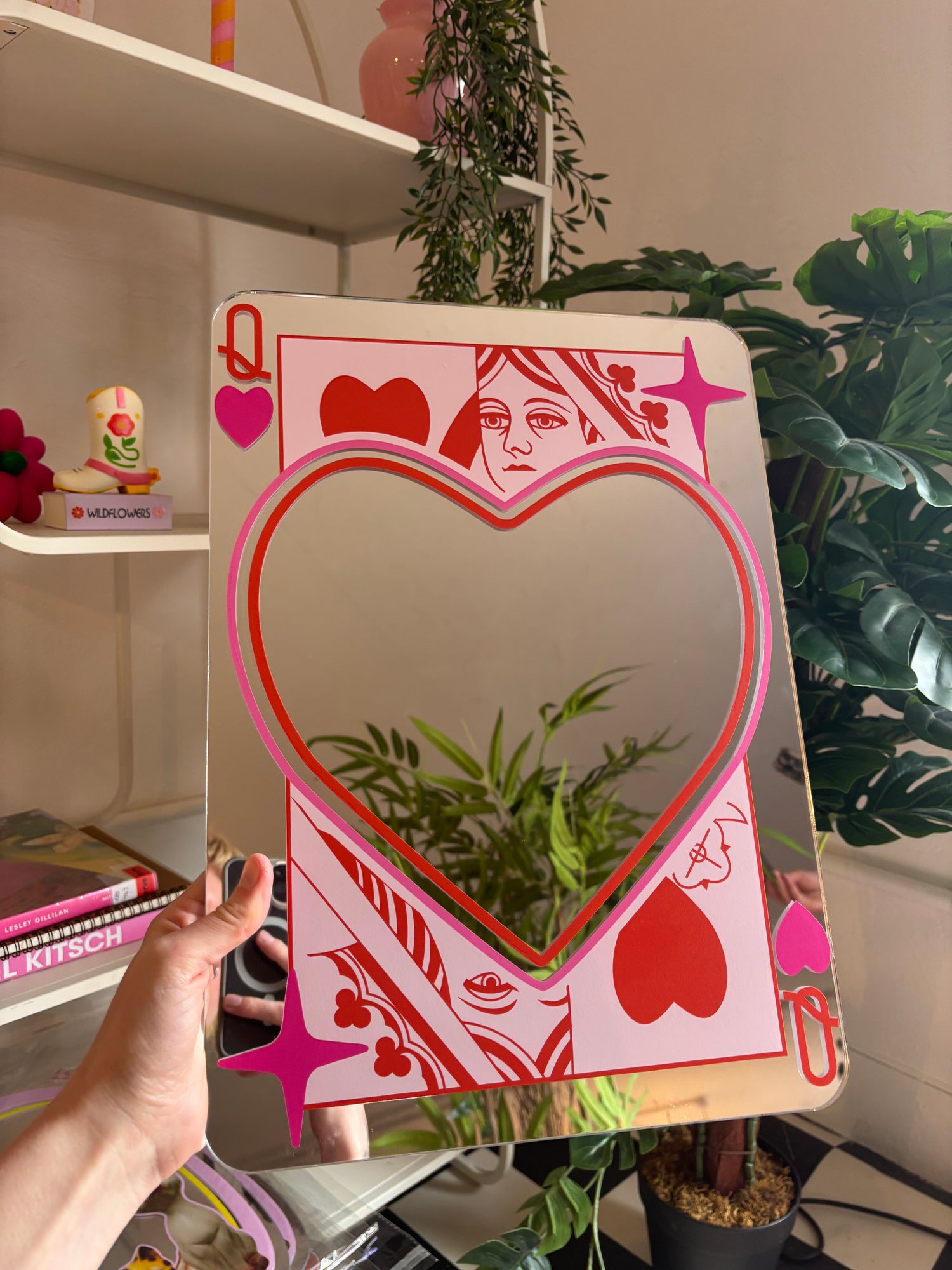 A hand is holding a large mirror with a heart-shaped design on it, surrounded by various plants and decorative items.