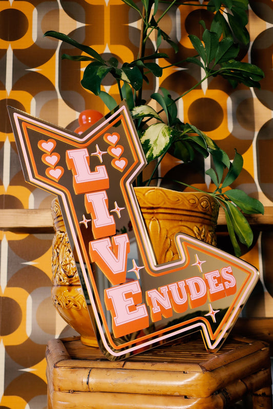 a wooden stool with a large, colorful sign that says "LIVE ENUDS" in bold, orange and pink letters. The sign is placed on top of the stool, and there is a potted plant nearby. The background has a patterned wallpaper with a retro, vintage style.
