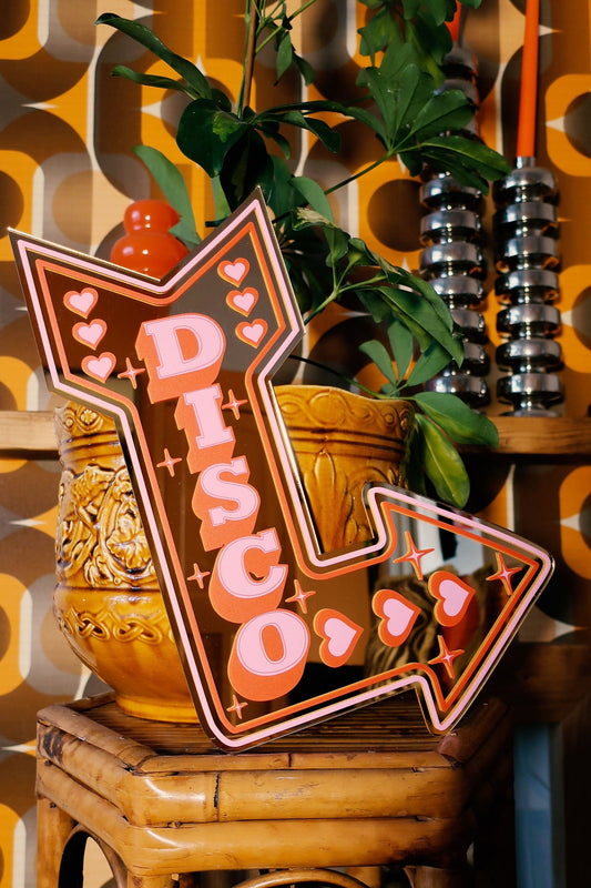 a vintage-style sign with a retro design, placed on a wooden stool in front of a plant and a shelf with decorative items.