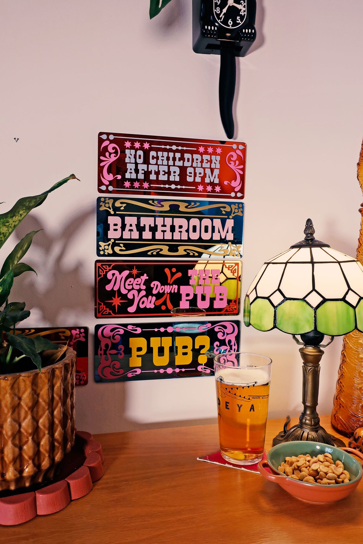a wooden table with a potted plant, a lamp, a glass of beer, and a bowl of snacks. There are also colorful signs on the wall, including one that says "No Children After 9PM" and another that says "Bathroom Meet Down You Pub".