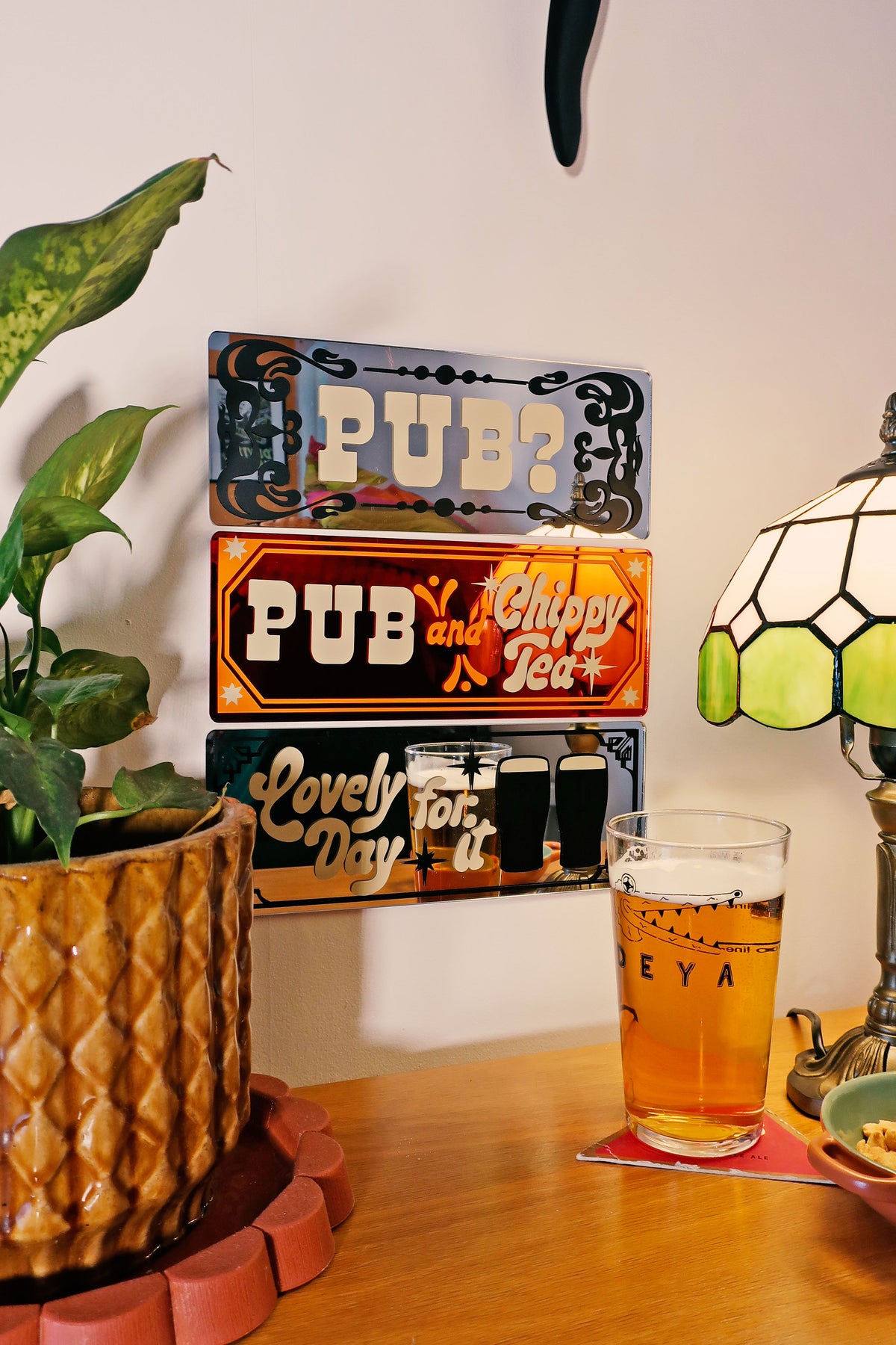 a wooden table with a potted plant, a glass of beer, and a sign that reads "PUB?" and "Chippy Tea".
