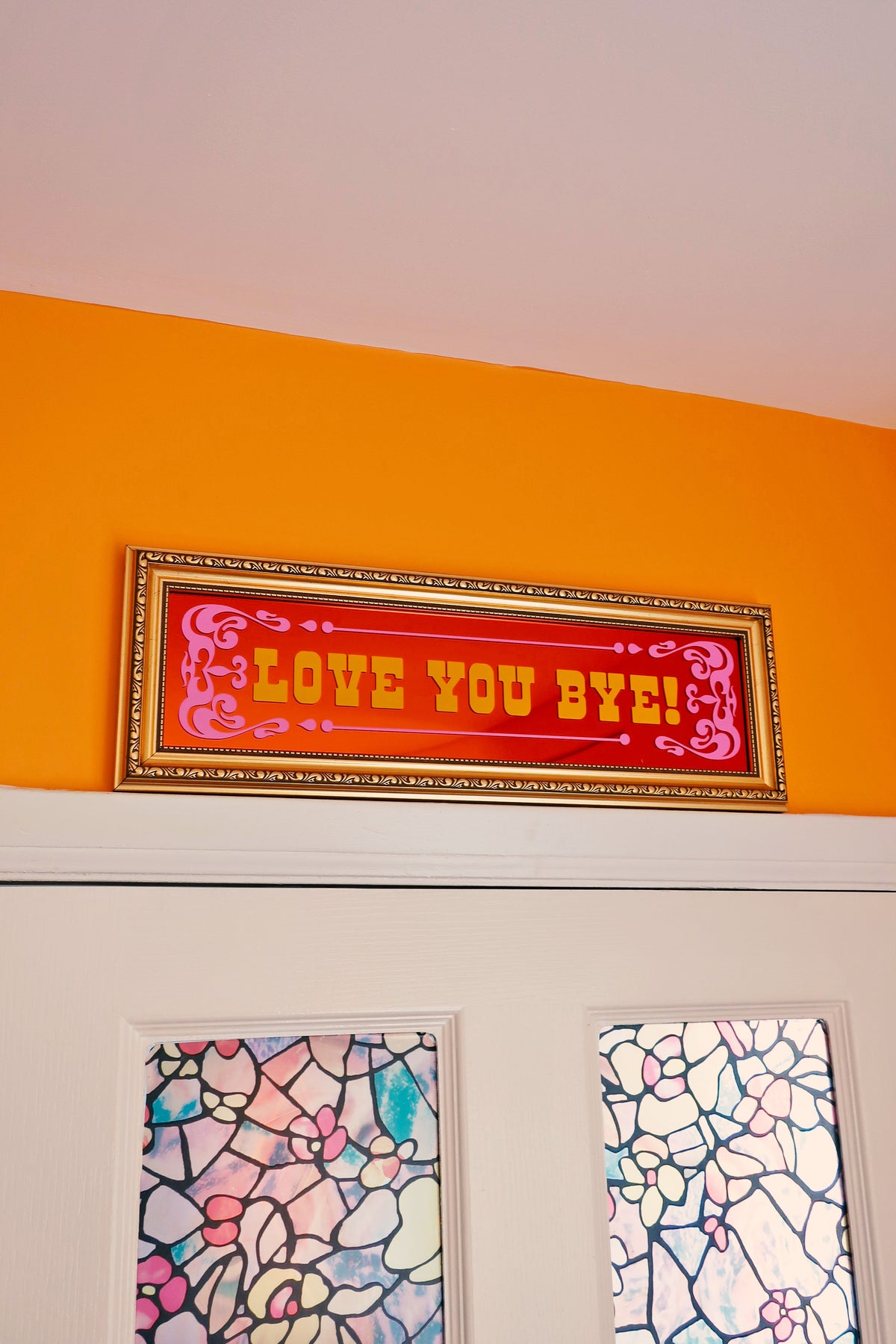 a framed sign with the text "LOVE YOU BYE" in a vibrant red and gold color scheme, hanging above a white door with a stained glass window.