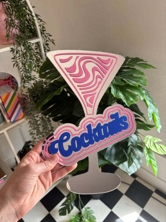 A hand is holding a pink and blue cocktail glass sign with the word "Cocktails" written on it, in front of a plant.