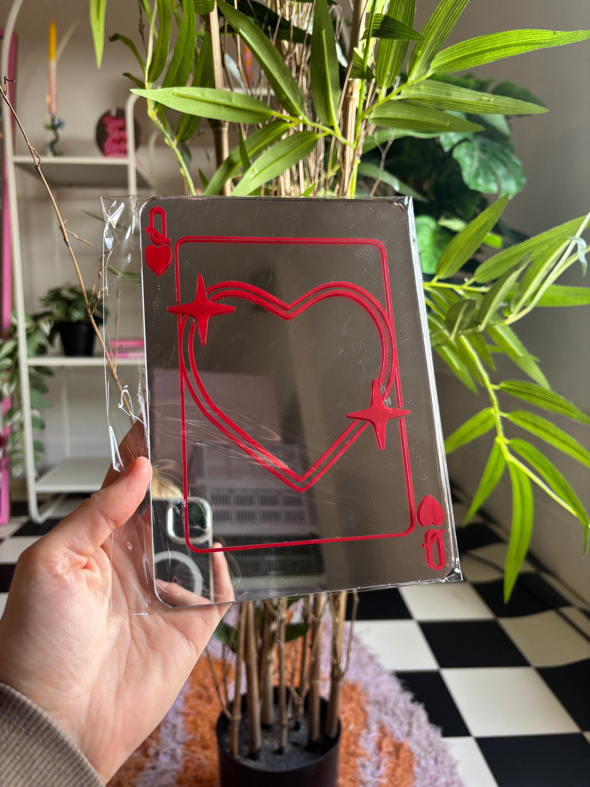 A hand is holding a mirror with a red heart and star design on it, in front of a potted plant.