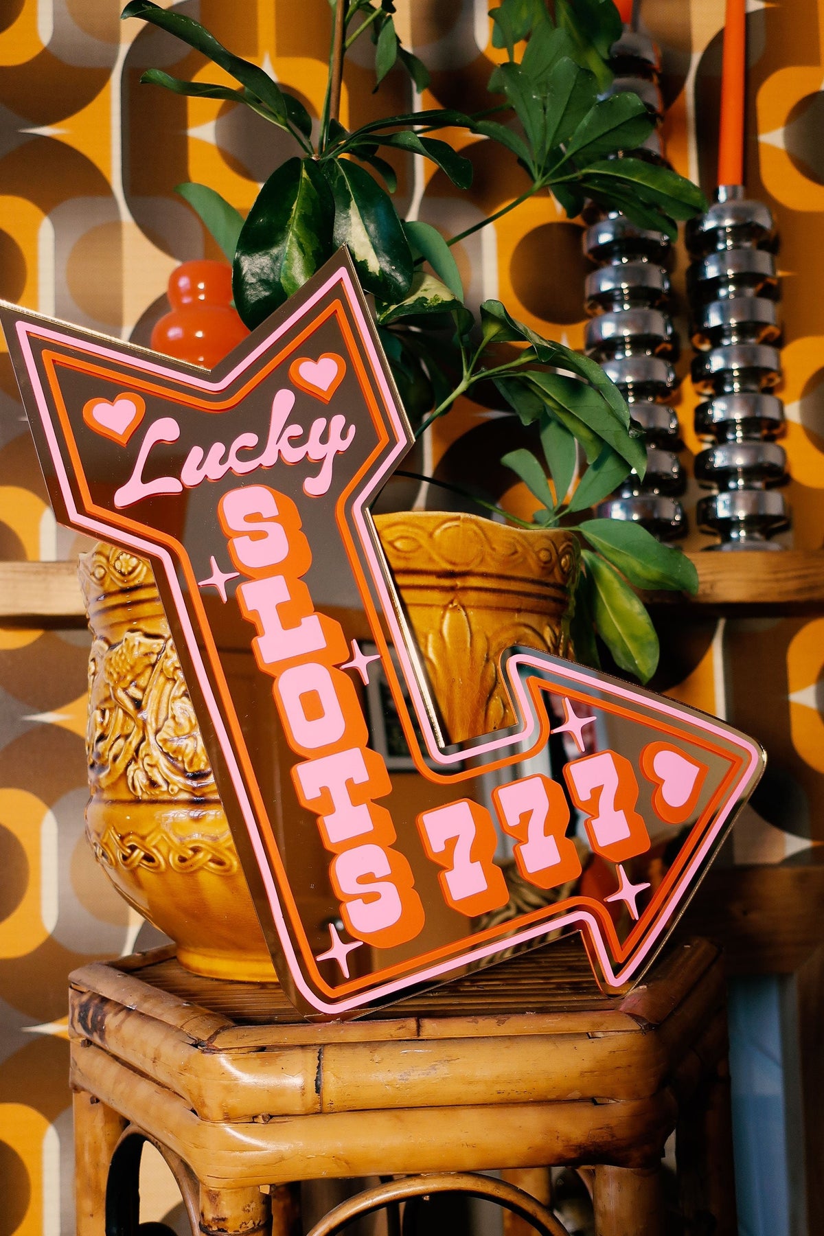 a wooden stool with a large, colorful sign that says "Lucky Stots 777" on it. The sign is placed on a wooden table, and there is a potted plant and some decorative items in the background.