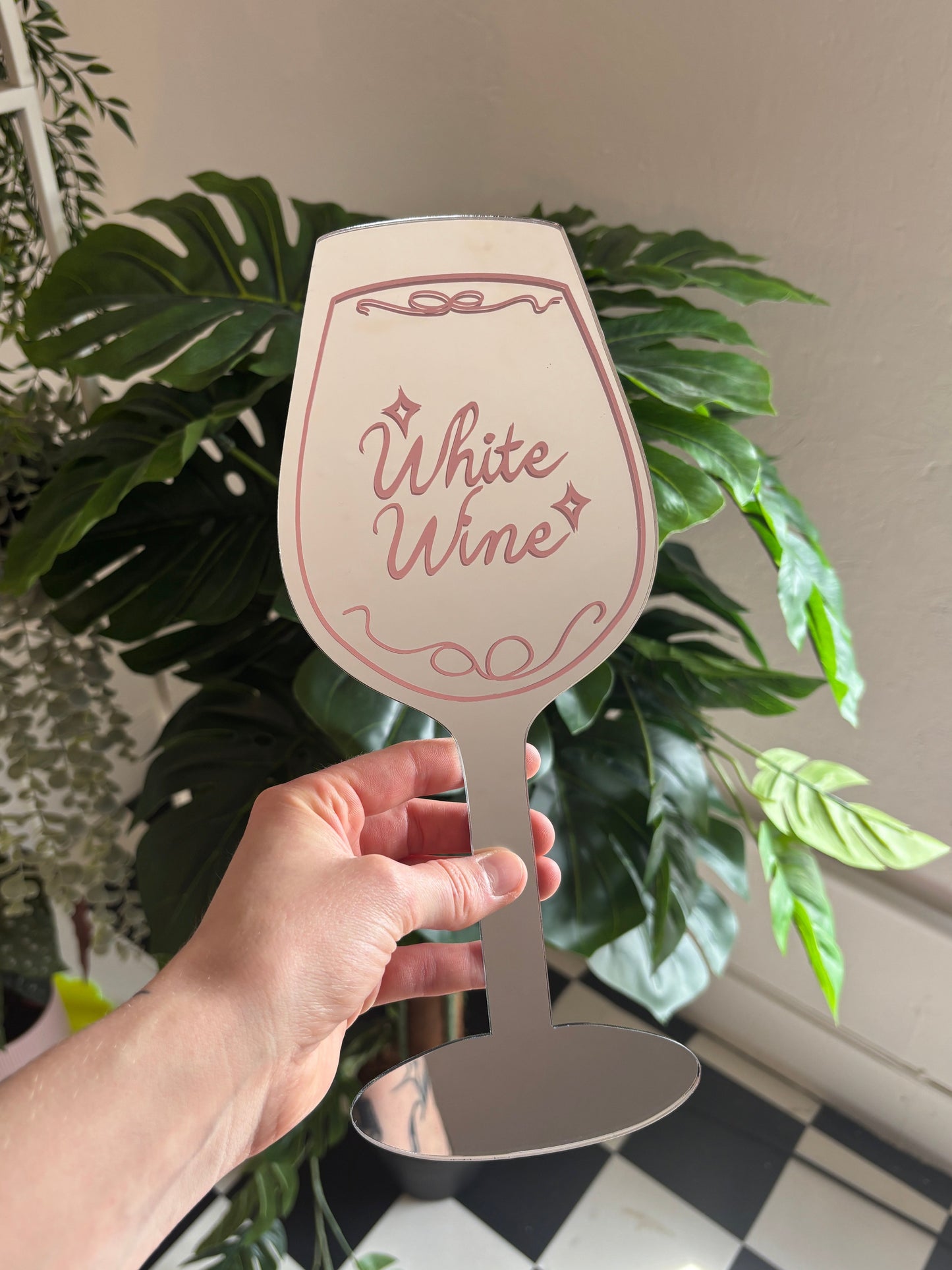 A hand holding a white wine glass with the text "White Wine" written on it, surrounded by various potted plants.