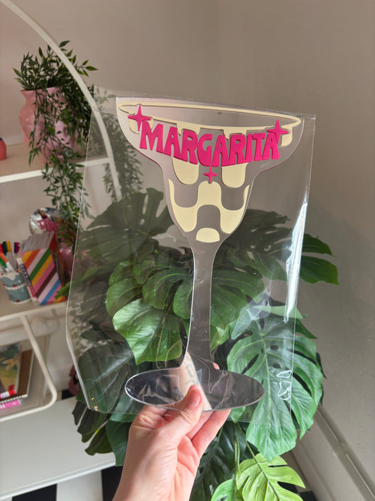 A hand is holding a clear plastic margarita glass with the word "MARGARITA" written on it.