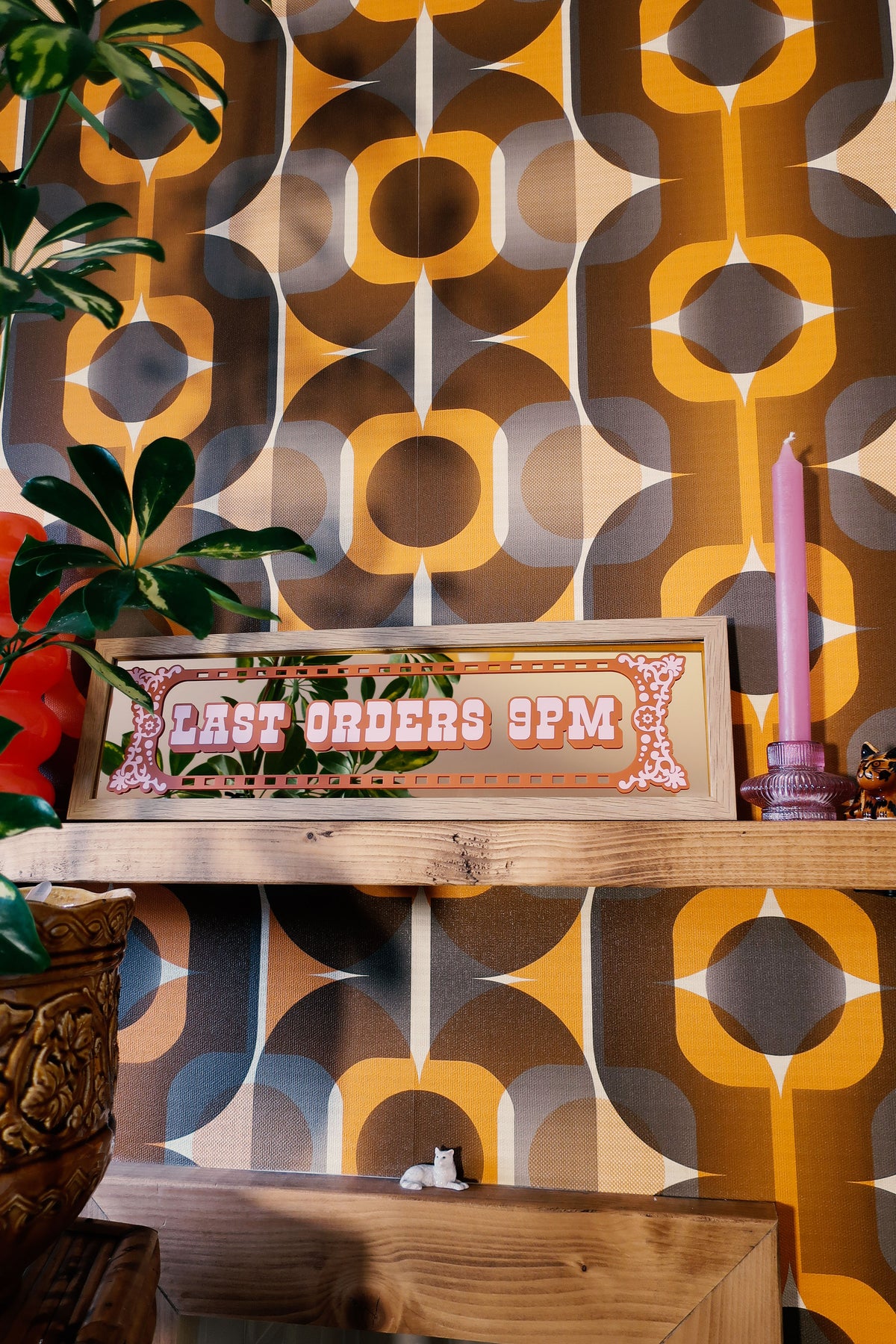 a shelf with a sign that reads "Last Orders 9PM", a pink candle, and a plant. The background has a patterned wallpaper with a repeating design of circles and lines in various shades of brown, orange, and yellow.