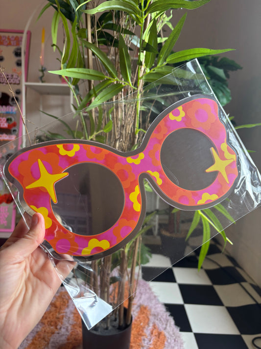 A hand is holding a colorful, patterned pair of sunglasses in front of a potted plant.