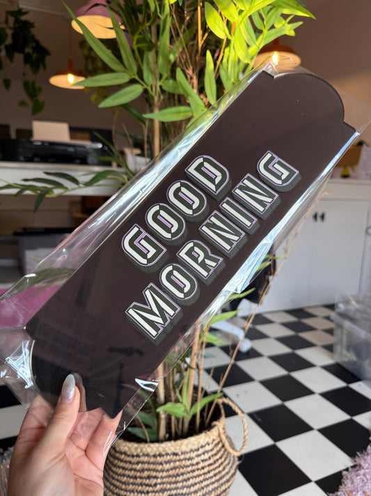 A person's hand holding a black sign with the text "GOOD MORNING" printed on it.