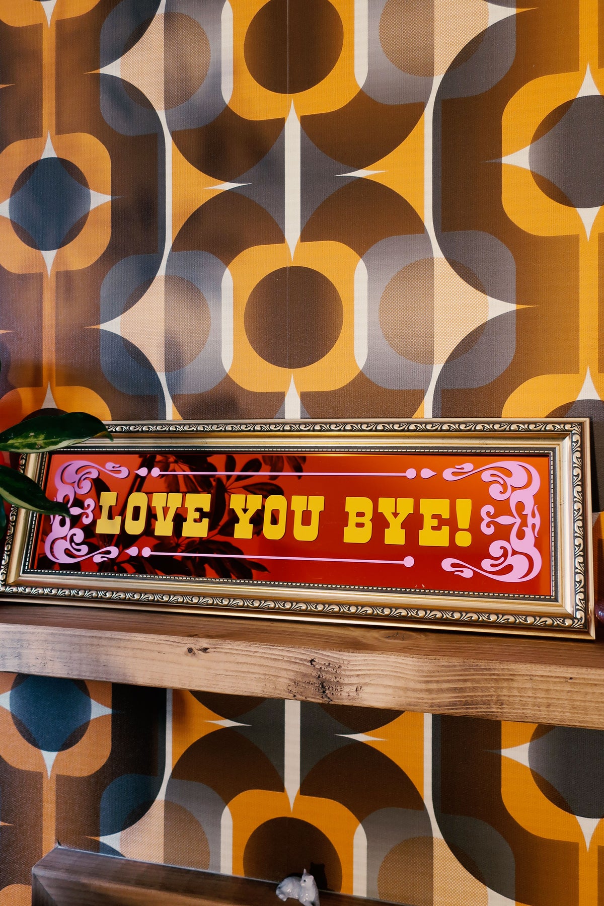 a framed sign with the text "LOVE YOU BYE!" displayed on a wooden shelf against a patterned wall.