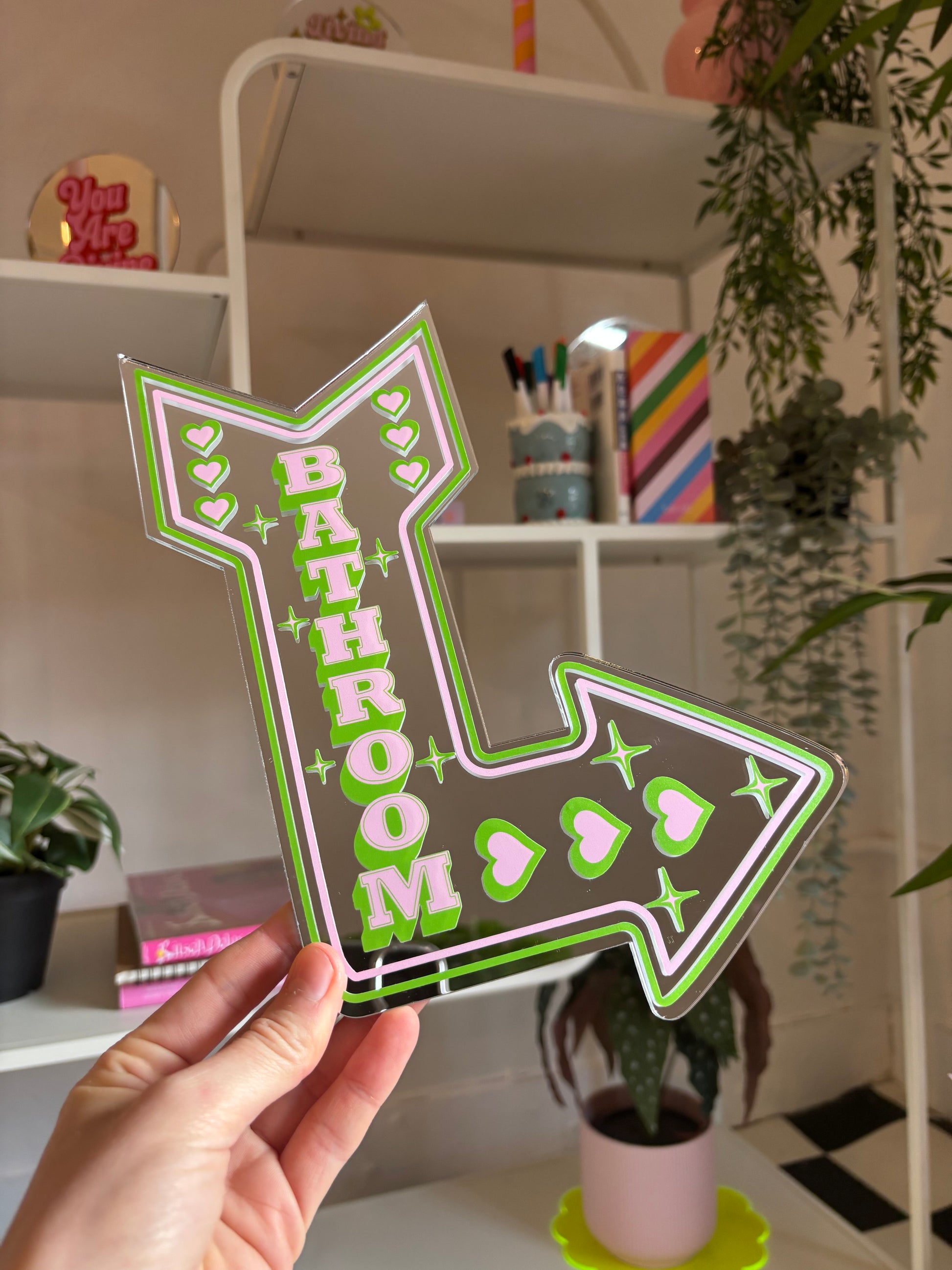 A hand is holding a large, colorful sign that says "BATHROOM" in a green and pink design, with stars and hearts around it.