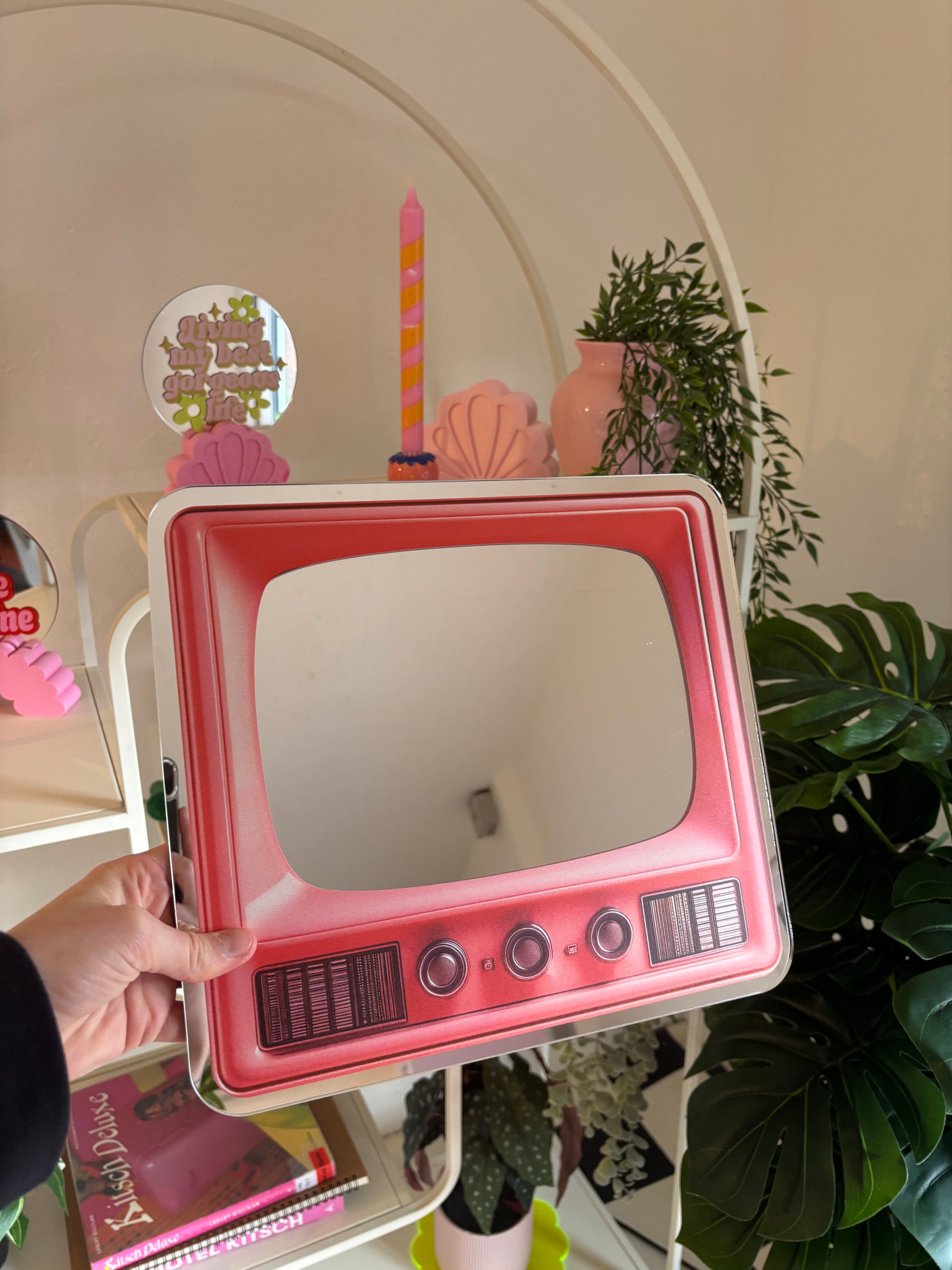 A hand is holding a pink, retro-style television set mirror in front of a white shelf with various decorative items, including a candle, a vase, and a potted plant.