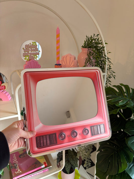 A hand is holding a pink, retro-style television set mirror in front of a white shelf with various decorative items, including a candle, a vase, and a potted plant.
