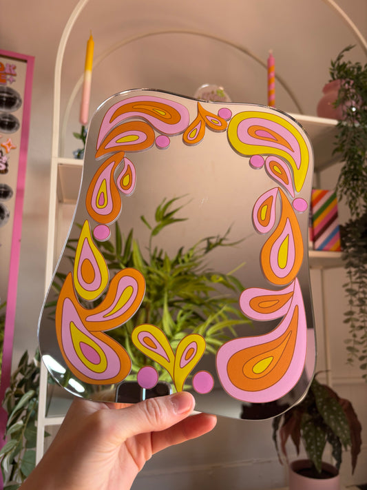 A hand is holding up a mirror with a colorful, abstract design featuring pink, orange, and yellow shapes.