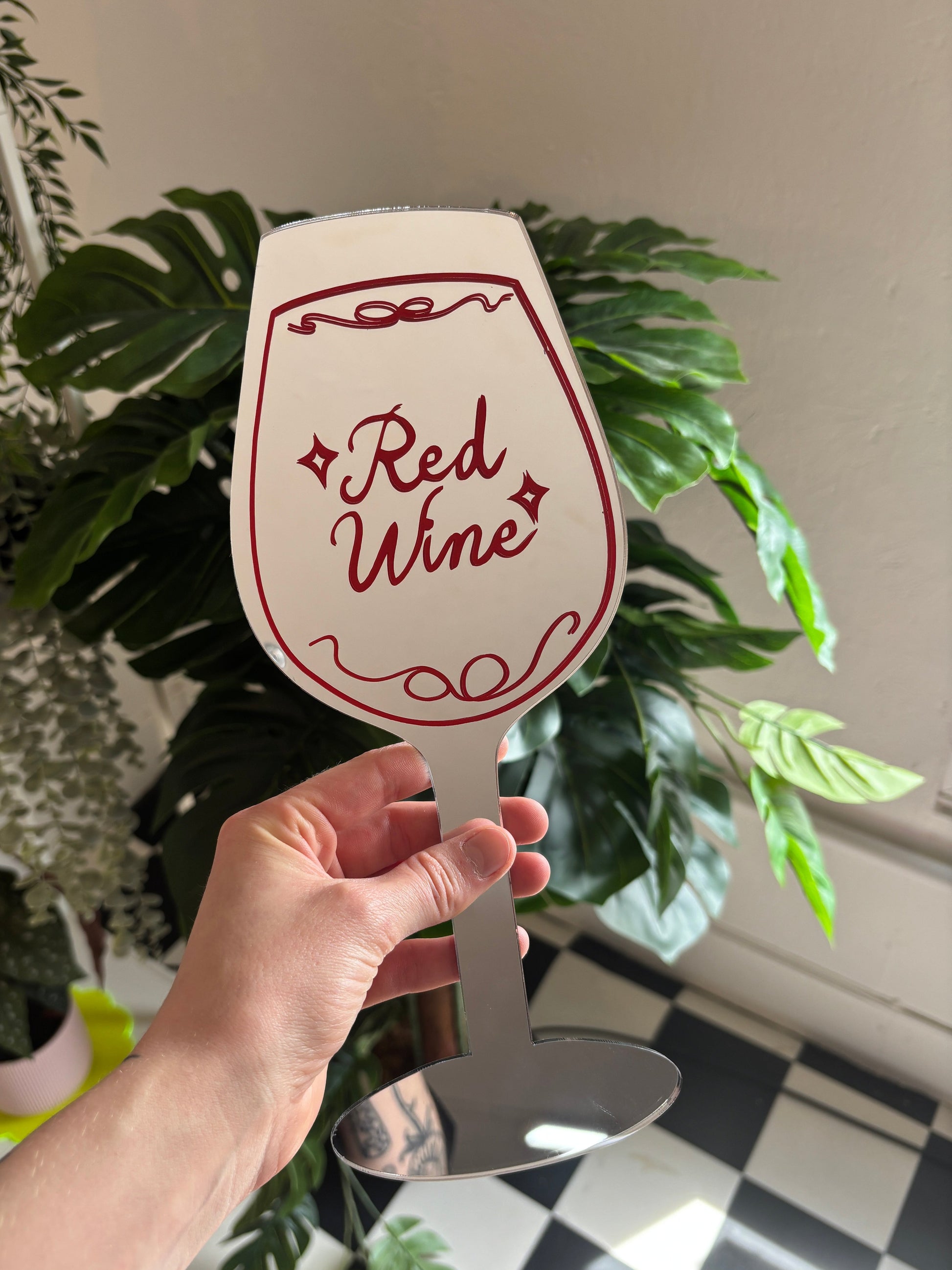 A hand holding a paper sign with the text "Red Wine" on it, surrounded by various plants and a checkered floor.