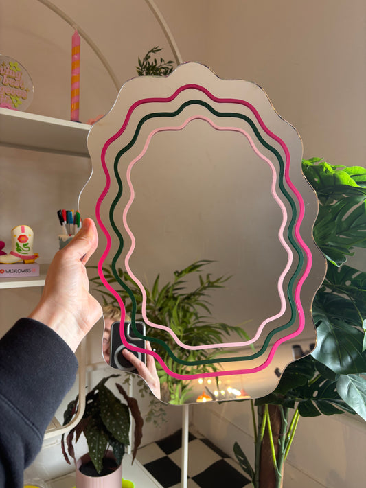 A hand is holding a colorful, wavy-shaped mirror in front of a shelf with various items, including a candle and plants.
