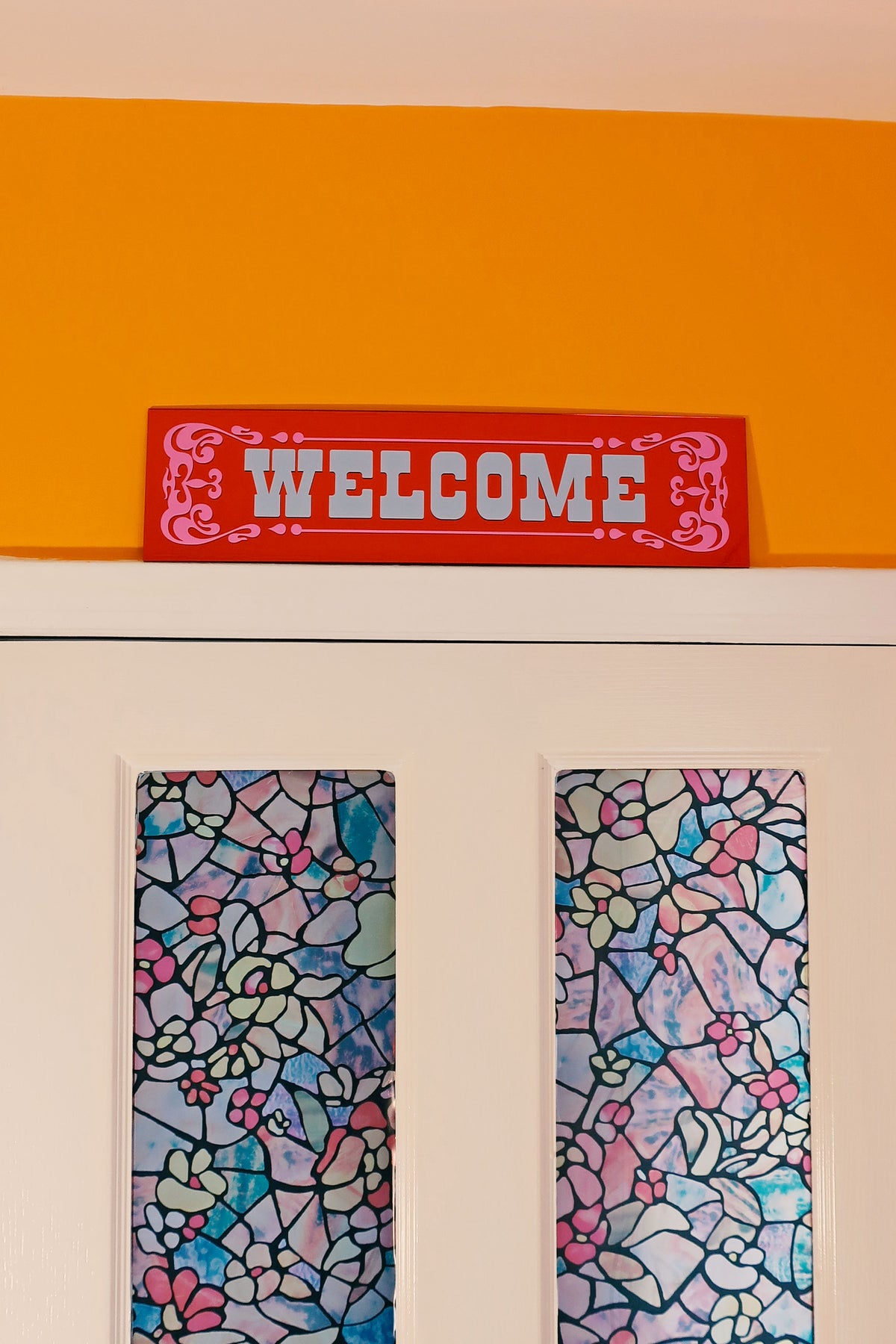 a red "Welcome" sign above a pair of stained glass windows with colorful floral patterns.