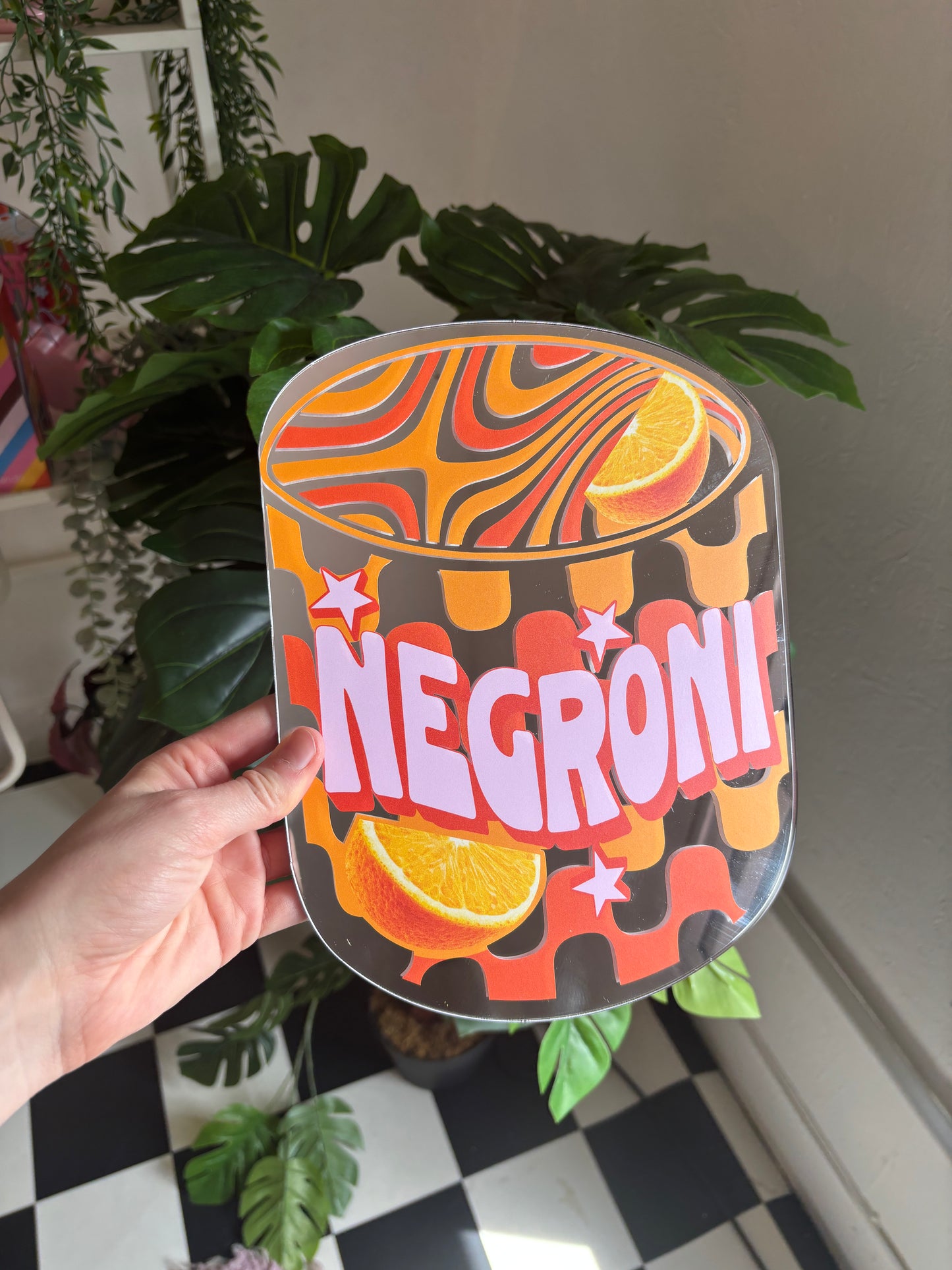 A hand is holding a colorful, abstract-style sign with the word "NEGRONI" written on it, surrounded by various plants and a checkered floor.