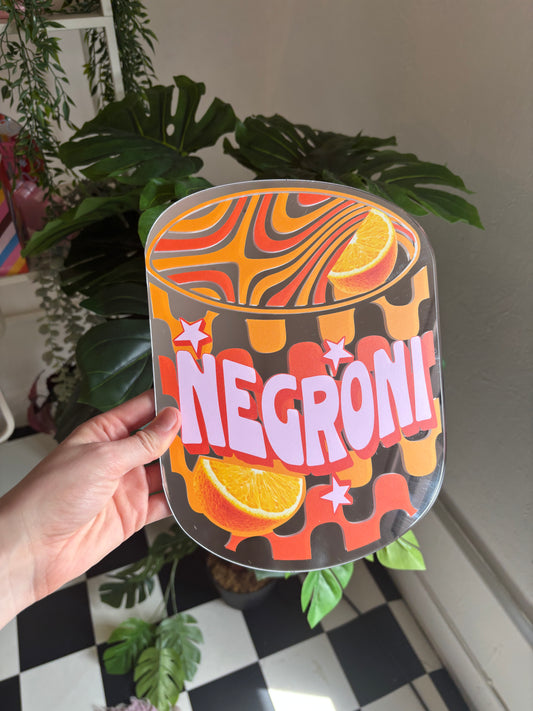 A hand is holding a colorful, abstract-style sign with the word "NEGRONI" written on it, surrounded by various plants and a checkered floor.
