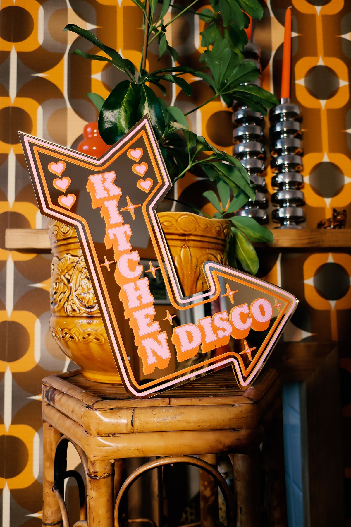 a sign that says "Kitchen Disco" in a decorative, stylized font, placed on a wooden stool. The background includes a patterned wall and a potted plant, creating a cozy and vibrant atmosphere.