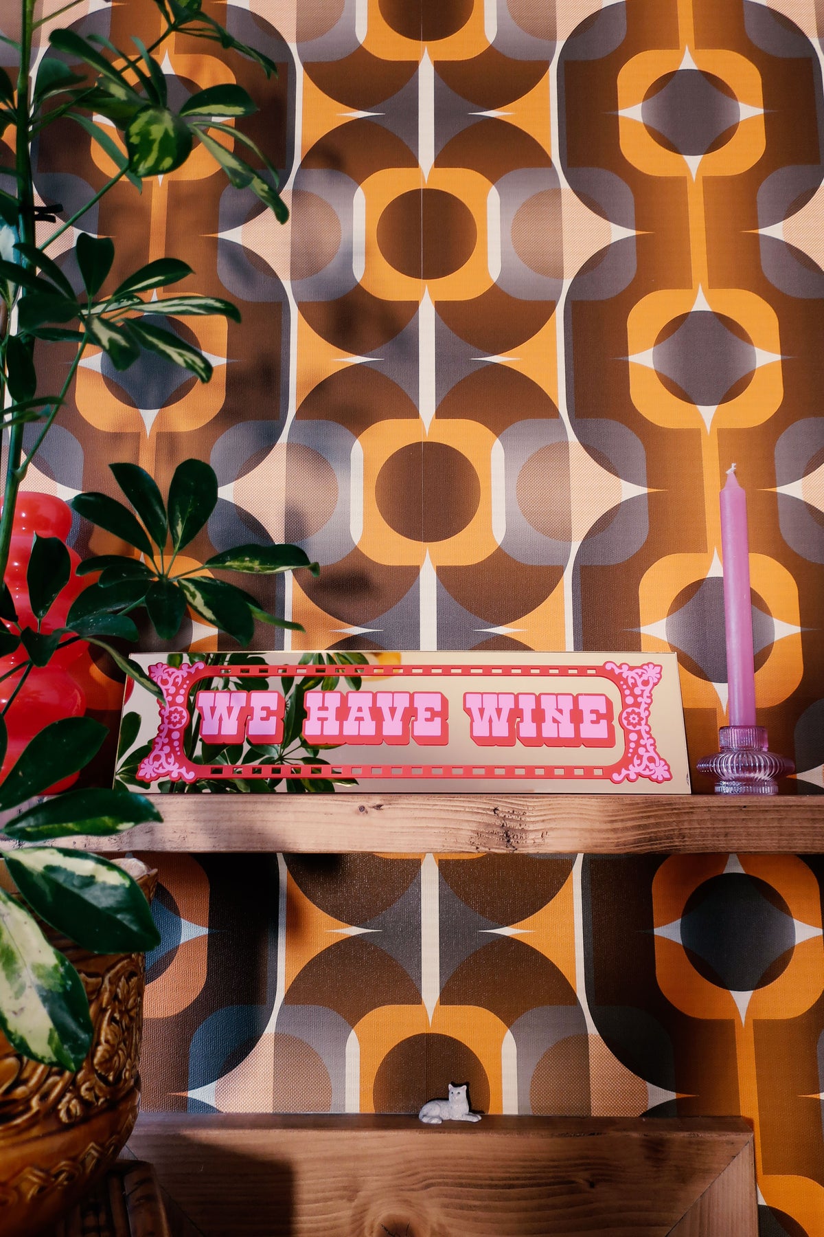 a shelf with a wooden frame, displaying a sign that reads "We have wine" in pink and red letters. The shelf is set against a wall with a repeating pattern of circles and lines in shades of orange, brown, and gray. A potted plant is also present in the scene.