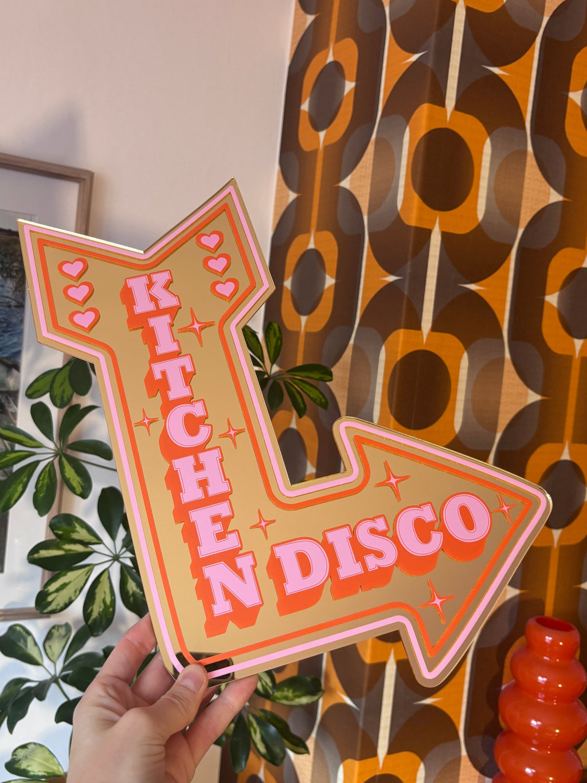 A hand is holding a large, colorful sign that says "Disco" in bold letters, with a patterned wall and plants in the background.