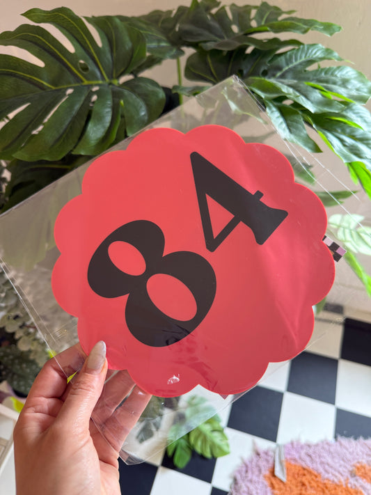 A hand is holding a clear plastic sheet with a red and black number 8 on it, against a backdrop of green plants and a checkered wall.
