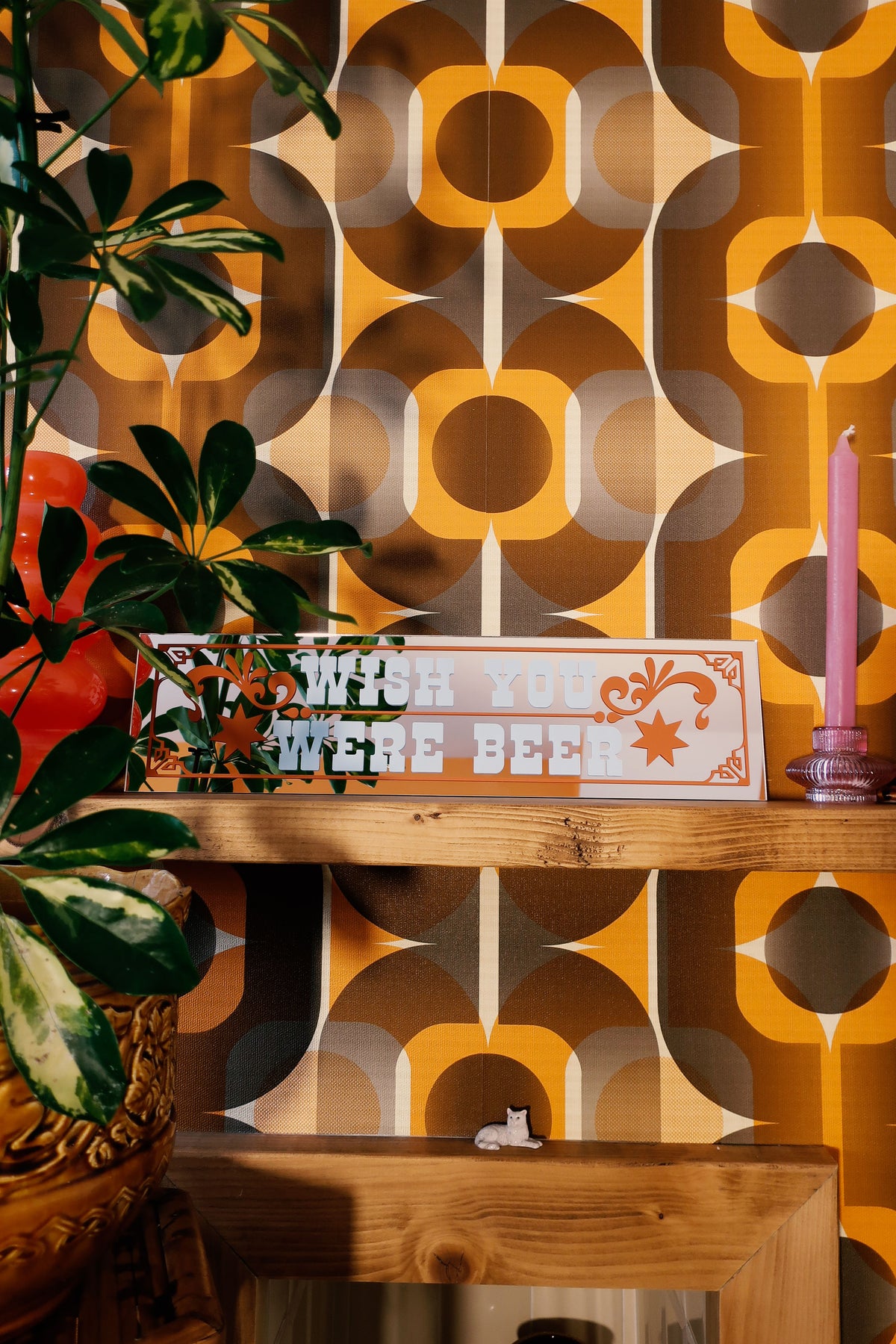 a wooden shelf with a sign that reads "Wish Your Beer Was Beer", a pink candle, and a potted plant. The background has a patterned wallpaper with a repeating design of circles and lines in shades of orange and brown.