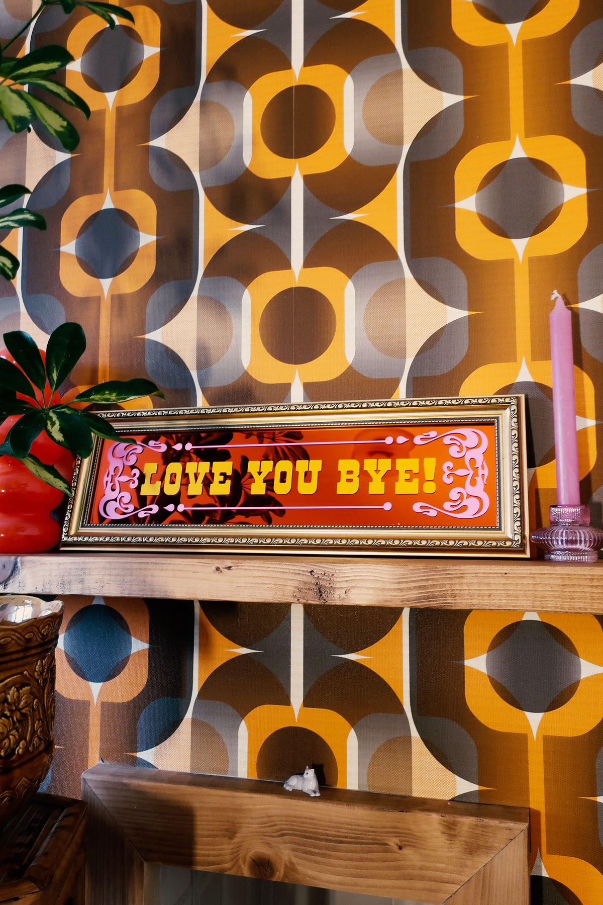 a framed sign with the text "LOVE YOU BYE!" displayed on a wooden shelf against a colorful, patterned wallpaper.