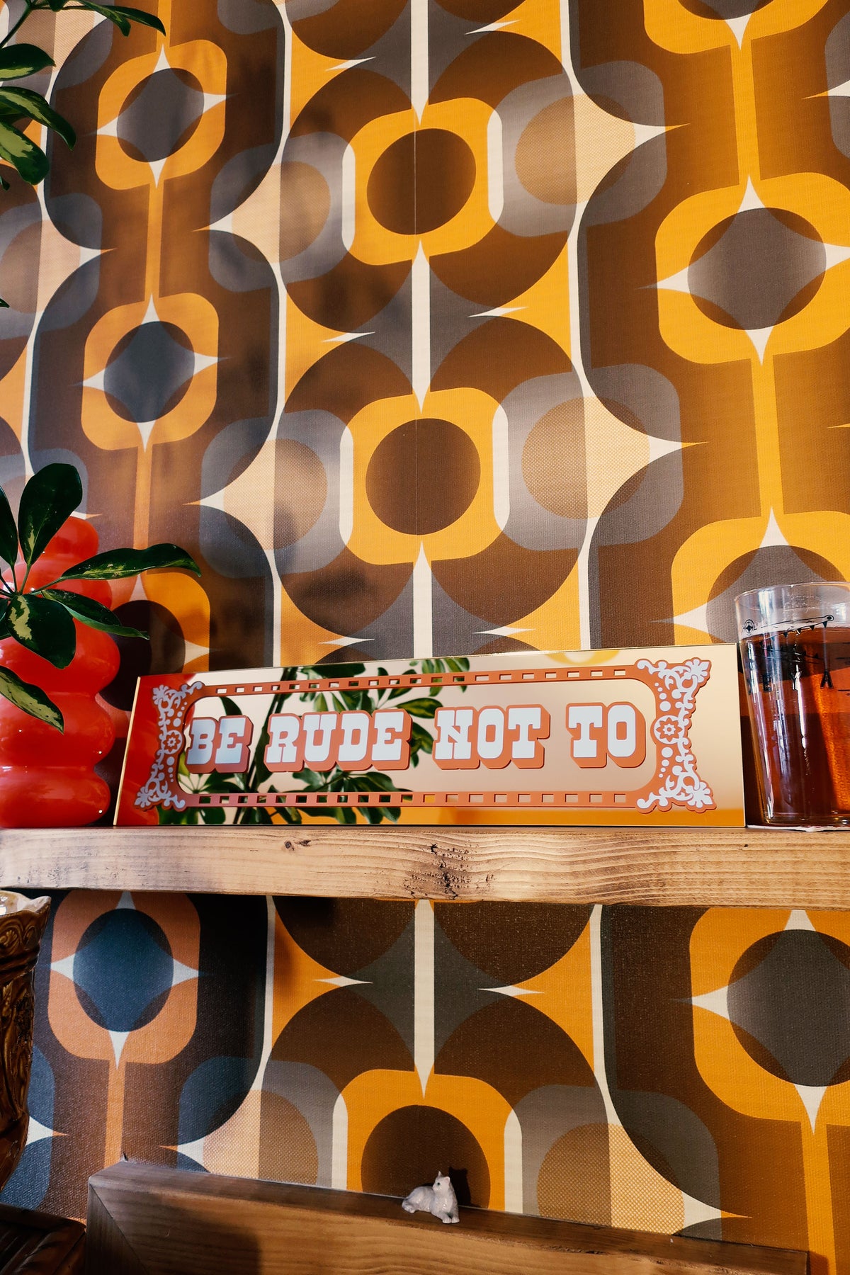 a wooden shelf with a sign that reads "Be Rude Not To", a glass of what appears to be a beverage, and a plant. The background has a patterned wallpaper with a repeating design of circles and lines in various shades of orange, brown, and gray.