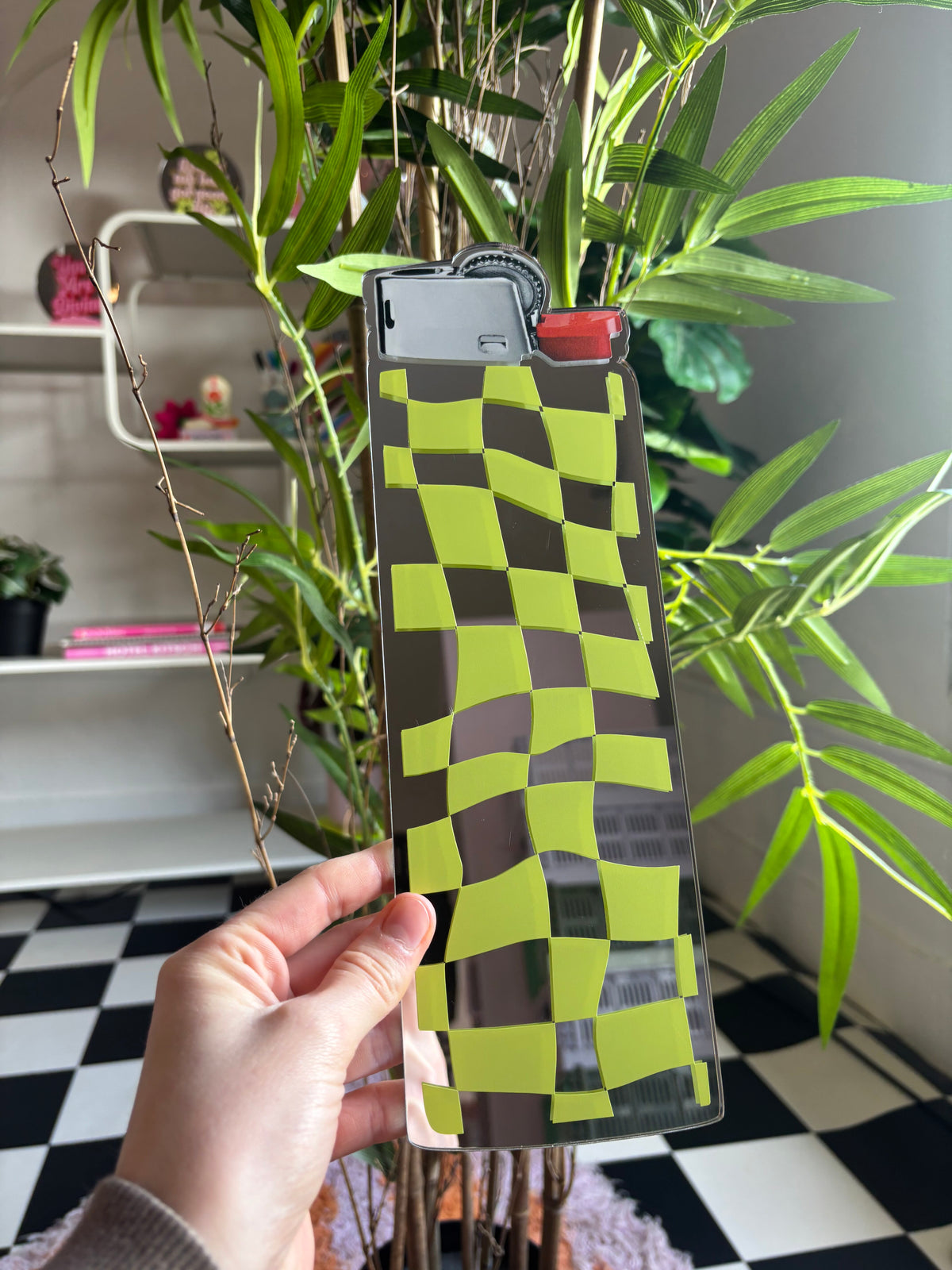 A hand is holding a large, checkered-patterned lighter in front of a plant with green leaves.