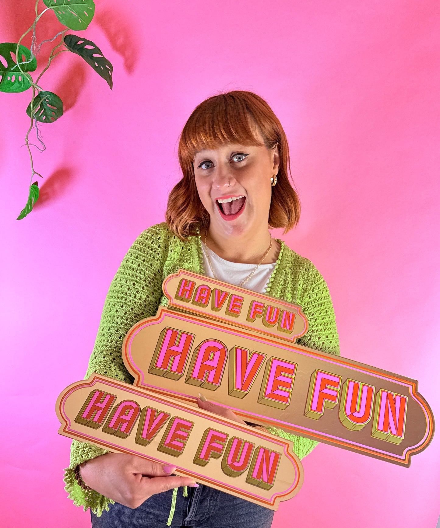 A woman in a green sweater is holding a sign that says "Have Fun" in front of a pink background.