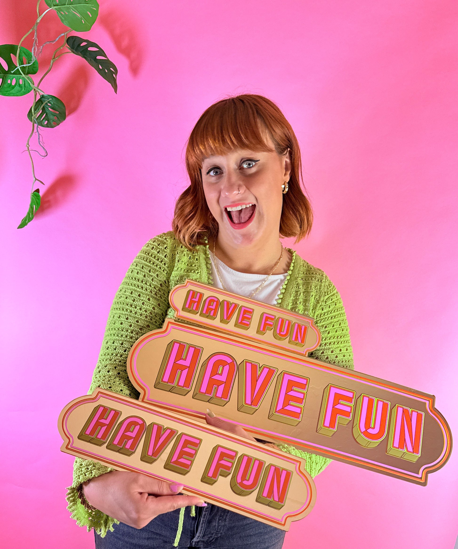 A woman in a green sweater is holding a sign that says "Have Fun" in front of a pink background.