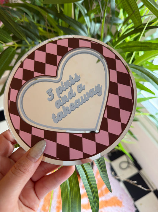 A hand holding a heart-shaped object with a checkered pattern and text on it, against a backdrop of green plants.