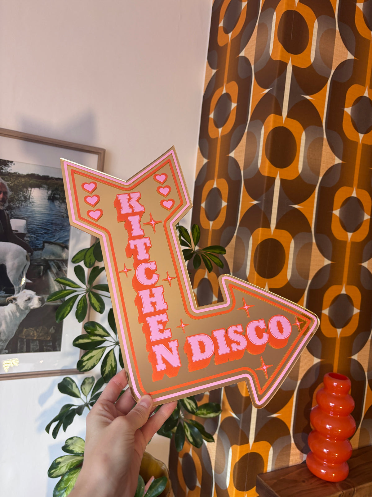 A hand is holding a sign that says "Disco Kitchen" in a room with a patterned wall and a plant.