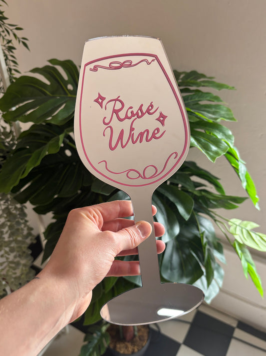 A hand holding a paper sign with the text "Roses and Wine" on it, surrounded by various potted plants.