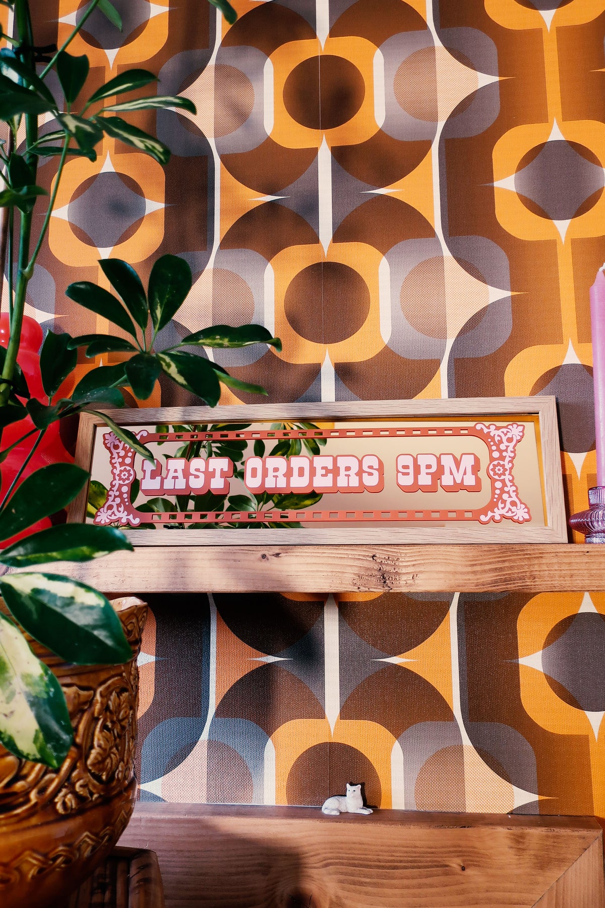 a wooden shelf with a plant and a sign that reads "LAST ORDER 9PM". The shelf is set against a wall with a patterned wallpaper.