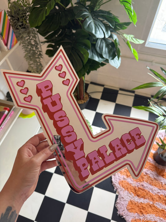 A hand holding a pink arrow-shaped sign with the word "PANIC" written on it, against a backdrop of various plants and a checkered floor.