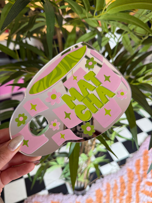 A hand holding a colorful, patterned object with the word "SHIT" written on it, surrounded by green leaves and a checkered background.