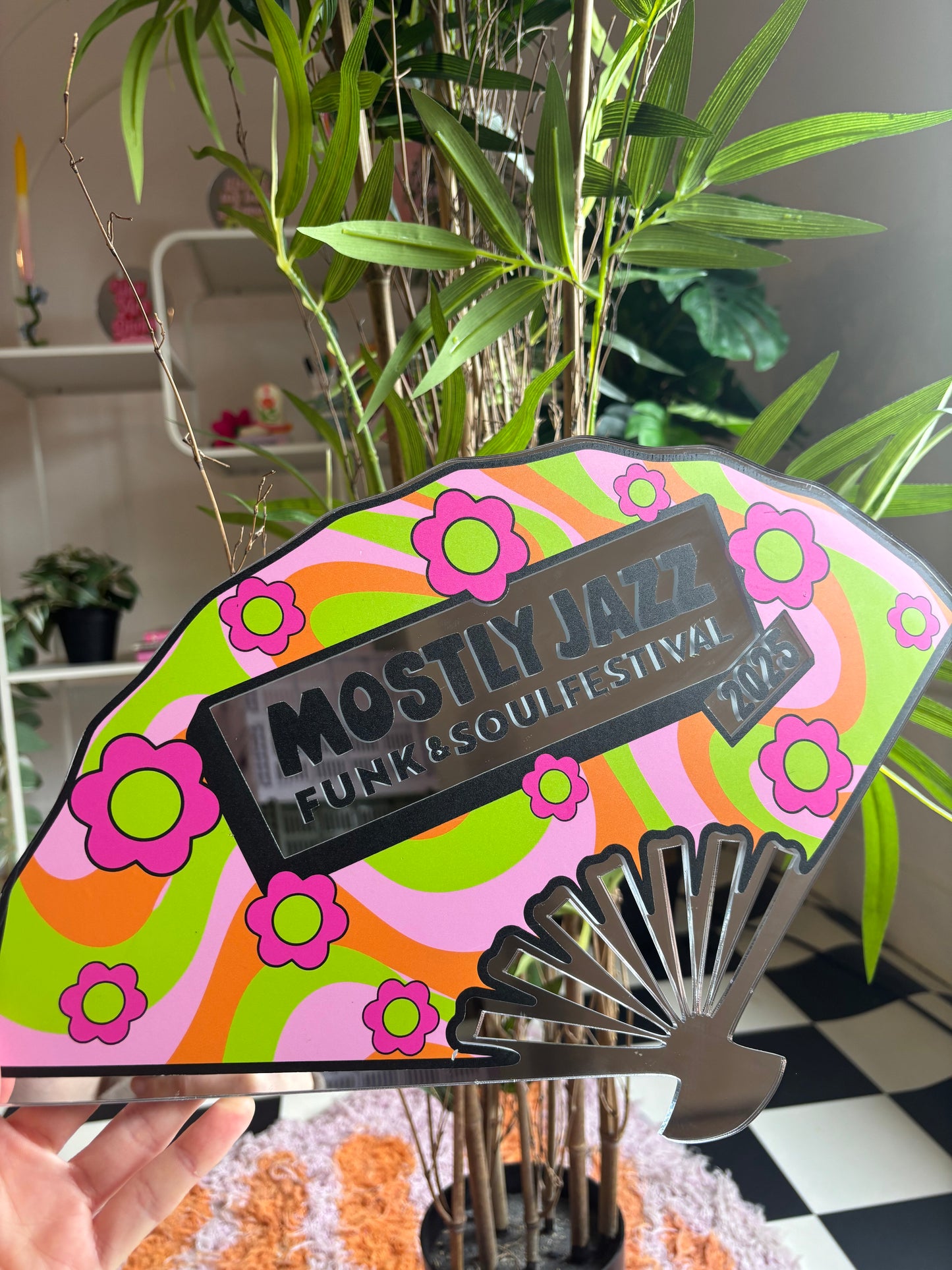 A colorful fan-shaped sign with the text "Mostly Jazz" and "Funk & Quilled Festival" printed on it, surrounded by a plant and a checkered floor.