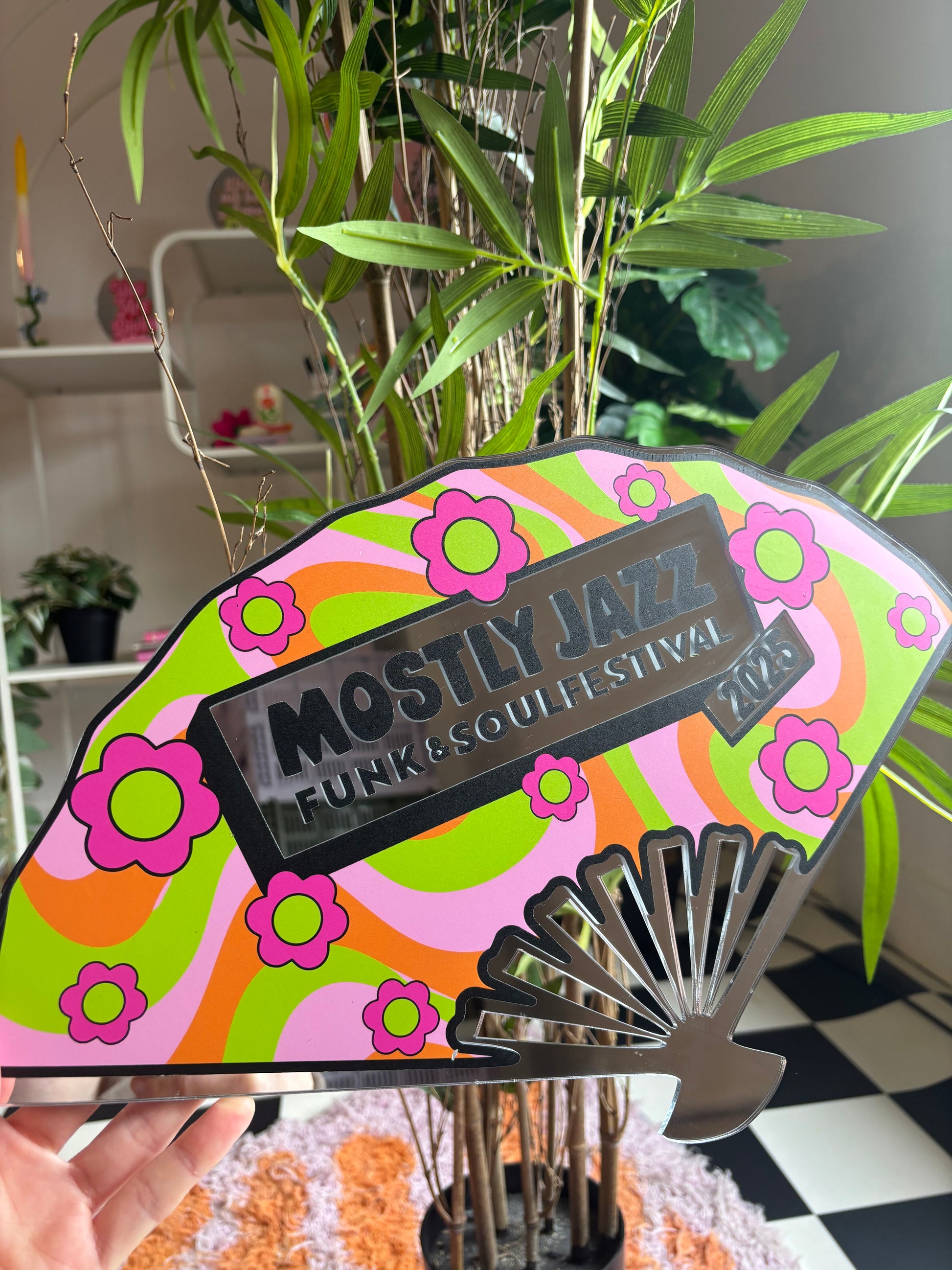 A colorful fan-shaped sign with the text "Mostly Jazz" and "Funk & Quilled Festival" printed on it, surrounded by a plant and a checkered floor.