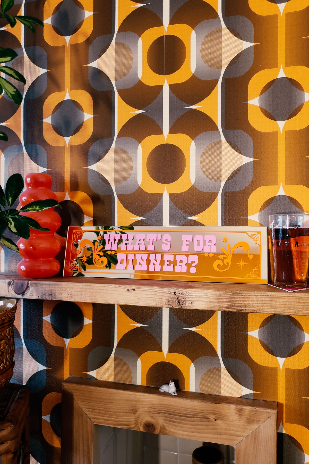 a wooden shelf with a sign that reads "What's for dinner?" and a glass of what appears to be a beverage. The background has a patterned wallpaper with a retro or vintage design.