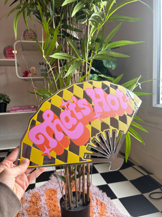 A hand is holding a colorful fan-shaped sign with the text "That's Hot" on it, which is placed in a potted plant.