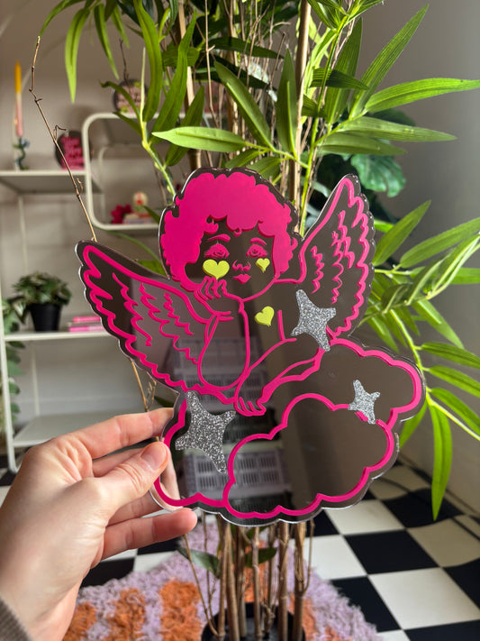 A hand is holding a colorful, stylized paper cutout of a cherub-like figure with wings and stars, set against a backdrop of green plants and a checkered floor.
